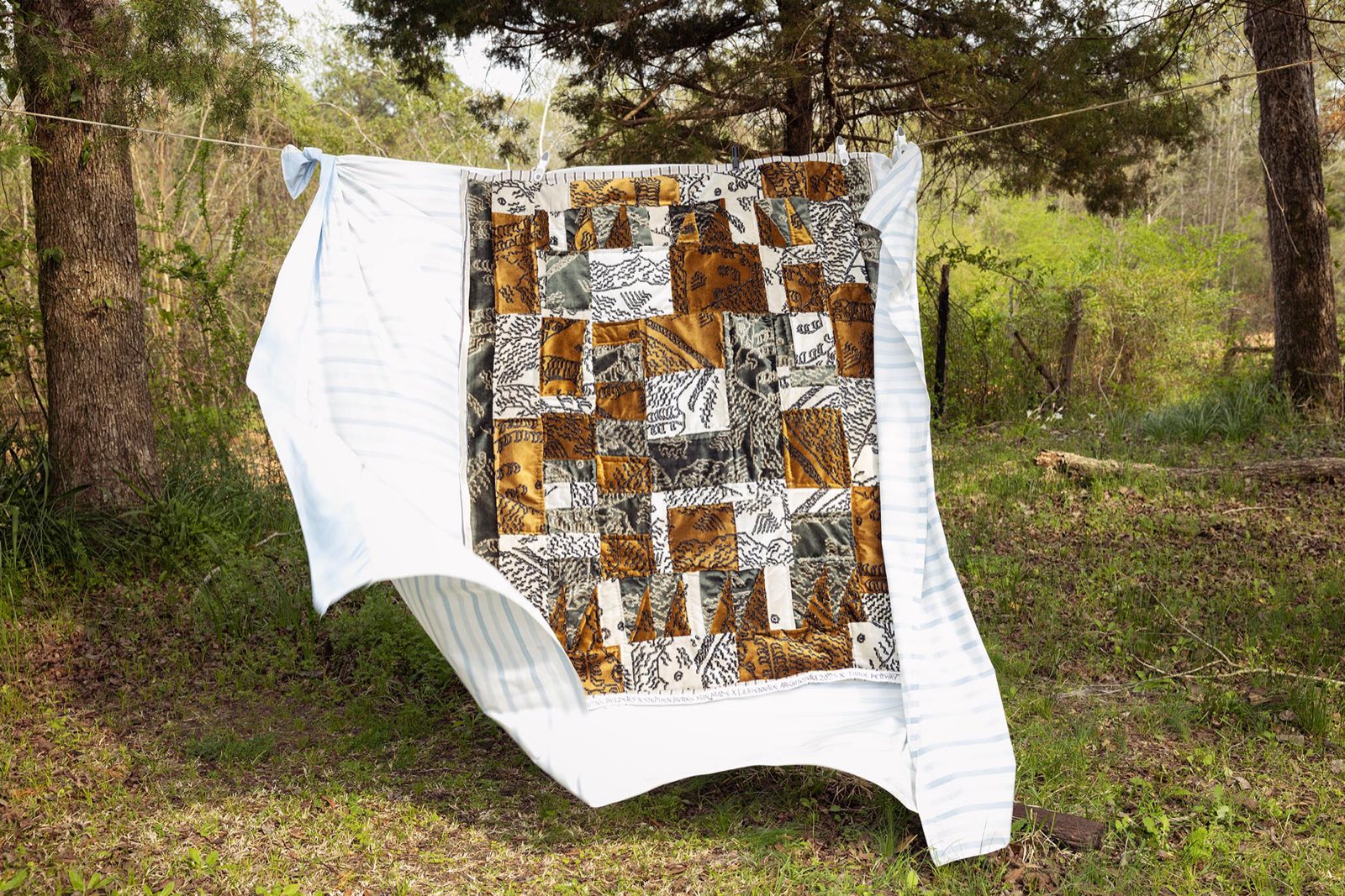 A Gee's Bend quilt in ochre, charcoal and cream Dedar Milano fabrics hanging on a clothesline between pine trees in Boykin, Alabama